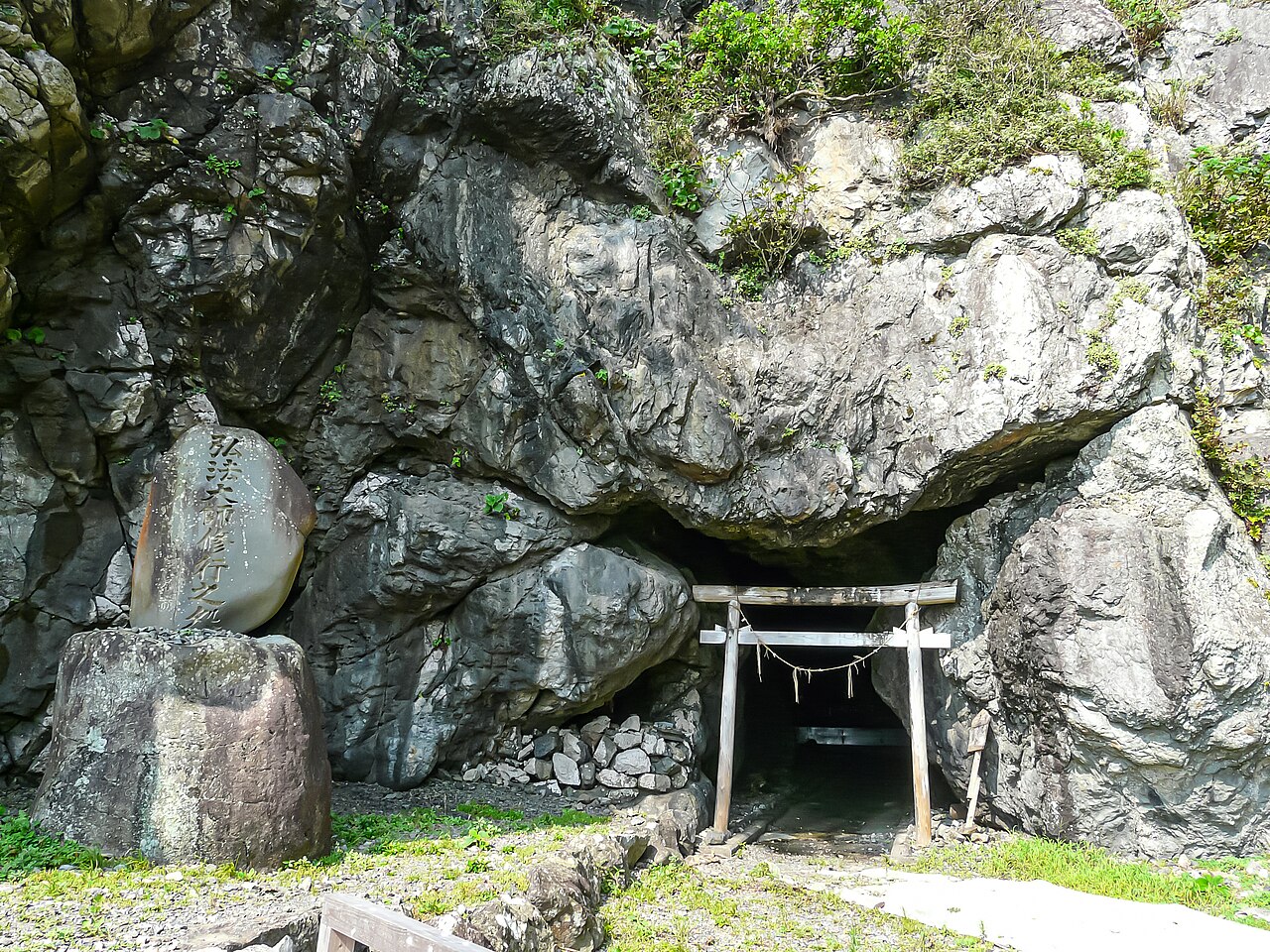 A small wooden torii standing at the mouth of a dark sea cave in a rock face, with a carved stone monument to Kobo Daishi's ascetic training at the left