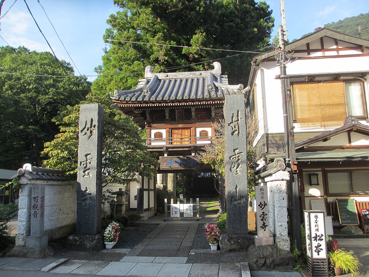 A tiled-roof temple gate building stands at the end of a paved approach lined with stone pillars, with trees behind it.