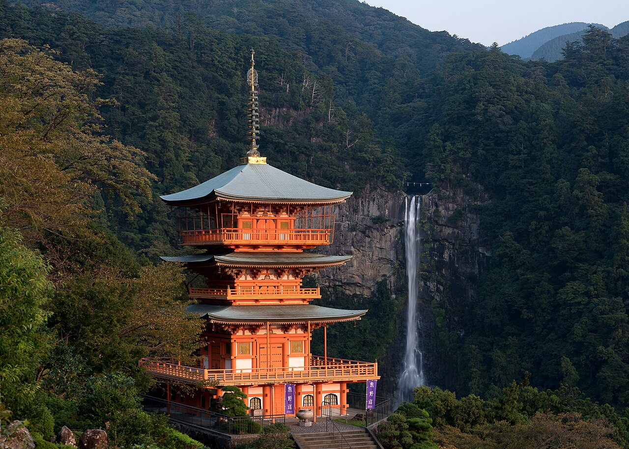 The vermilion three-storied pagoda of Seiganto-ji stands on a wooded hillside beside the sheer rock face of Nachi Falls, the waterfall's white ribbon visible in the distance under a dusky sky.