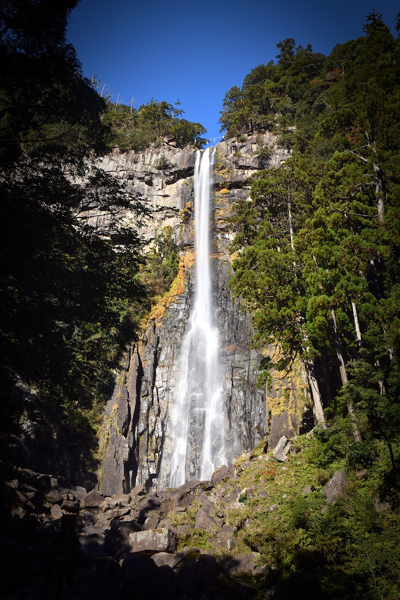 Nachi Falls drops as a single narrow ribbon of white water down a tall grey rock face between dense cedar-covered slopes, with a boulder-strewn streambed at its base under a clear blue sky.