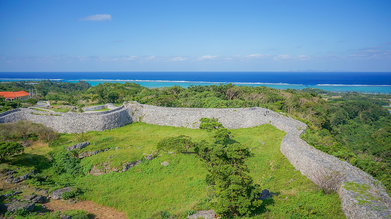 Curving stone walls of the Nakijin Castle ruins on a green ridge, with the blue sea and reef beyond