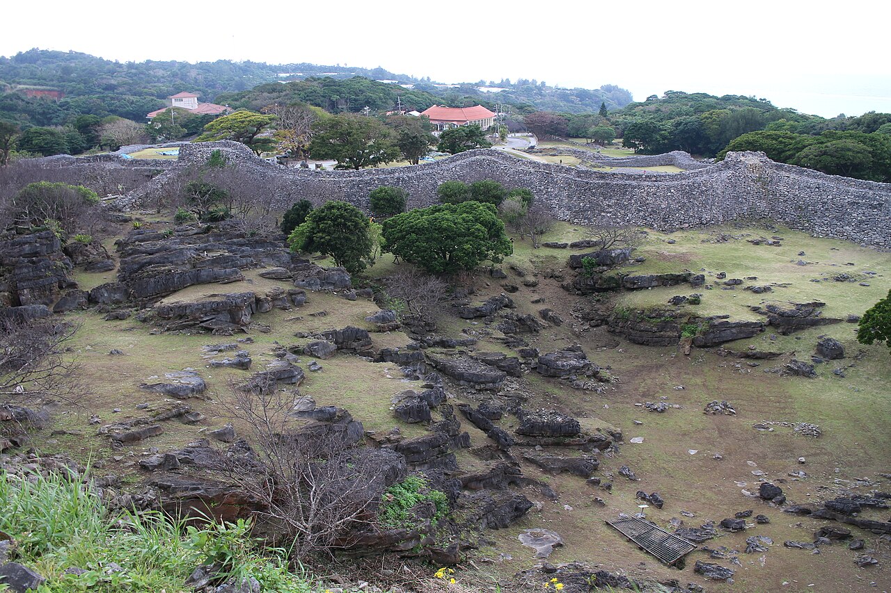 A long grey limestone wall of Nakijin Castle above the rocky ruins of the inner enclosures, with village rooftops behind