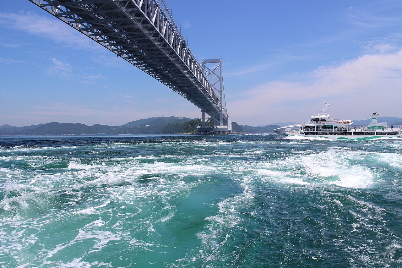 A white sightseeing boat crossing the churning green water of the Naruto Strait beneath the underside of the Onaruto Bridge, with a low hill in the distance.