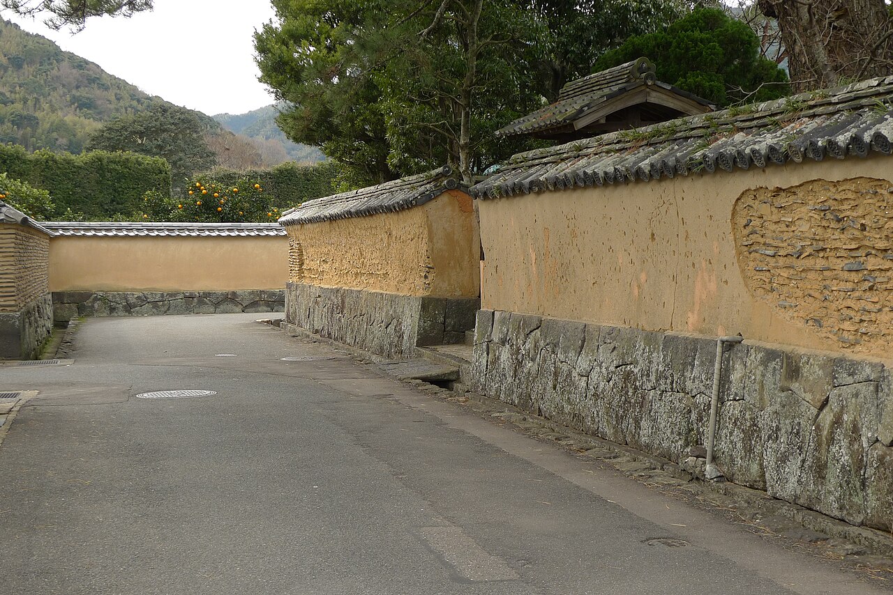 An empty lane in the Hiyako quarter of Hagi between ochre earthen walls on cut-stone bases, with orange citrus fruit showing above the wall and wooded hills behind