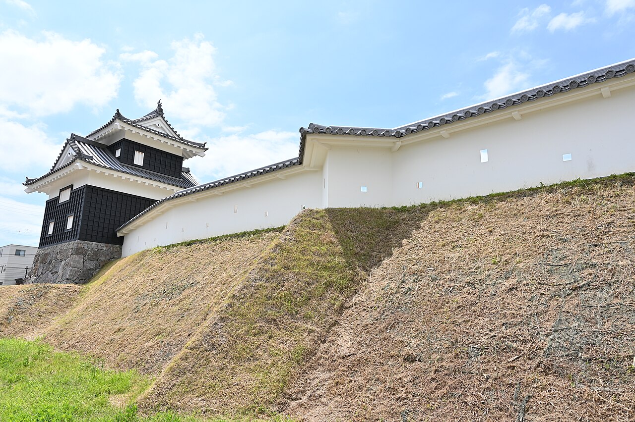 The Ushitora corner turret of Nishio Castle above a white-plastered wall and a grassy stone-faced bank under a blue sky