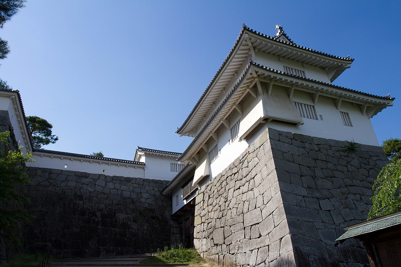 A white two-storey castle gate with tiled eaves seen from below, rising above a tall dry-laid stone wall under a clear blue sky
