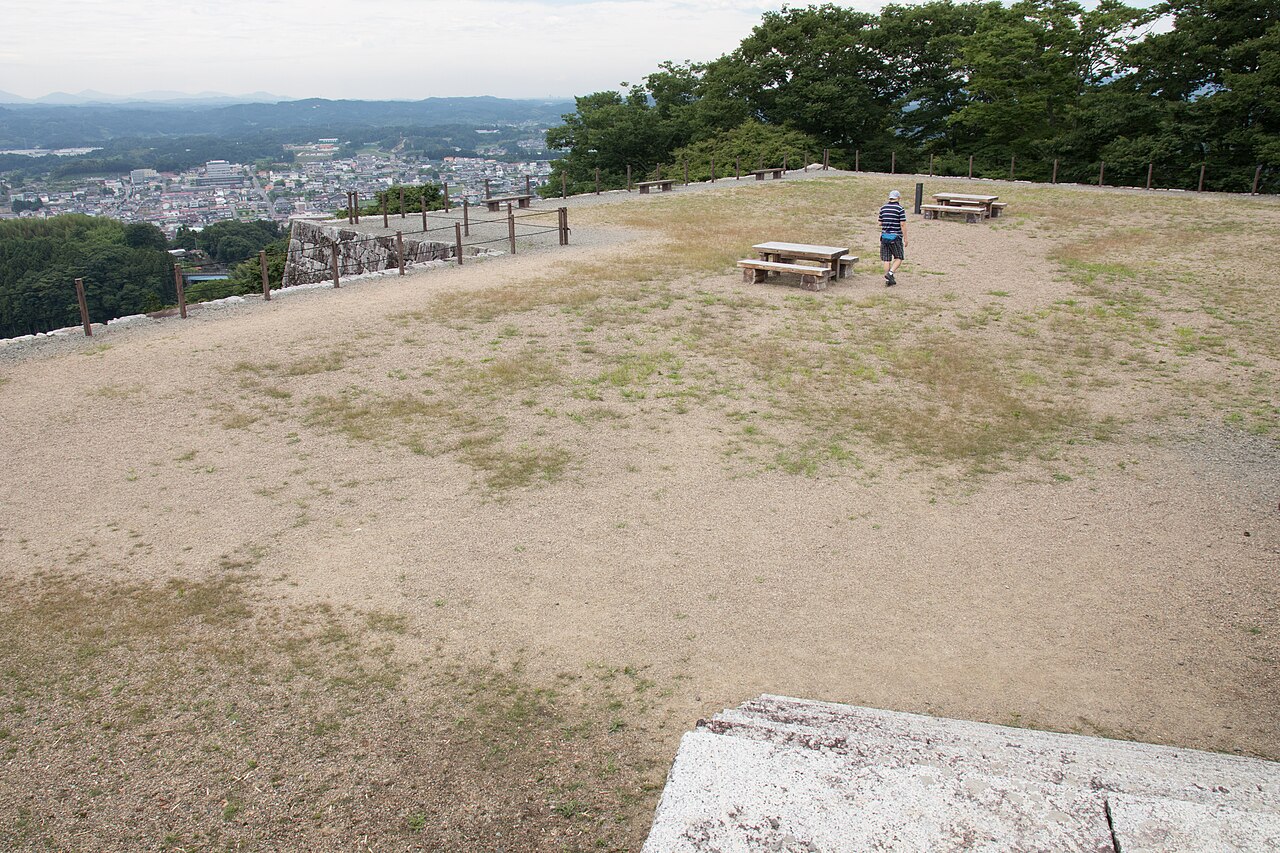 A wide gravel clearing on a hilltop castle site with picnic benches, a low stone wall at the edge and a town spread out in the valley below