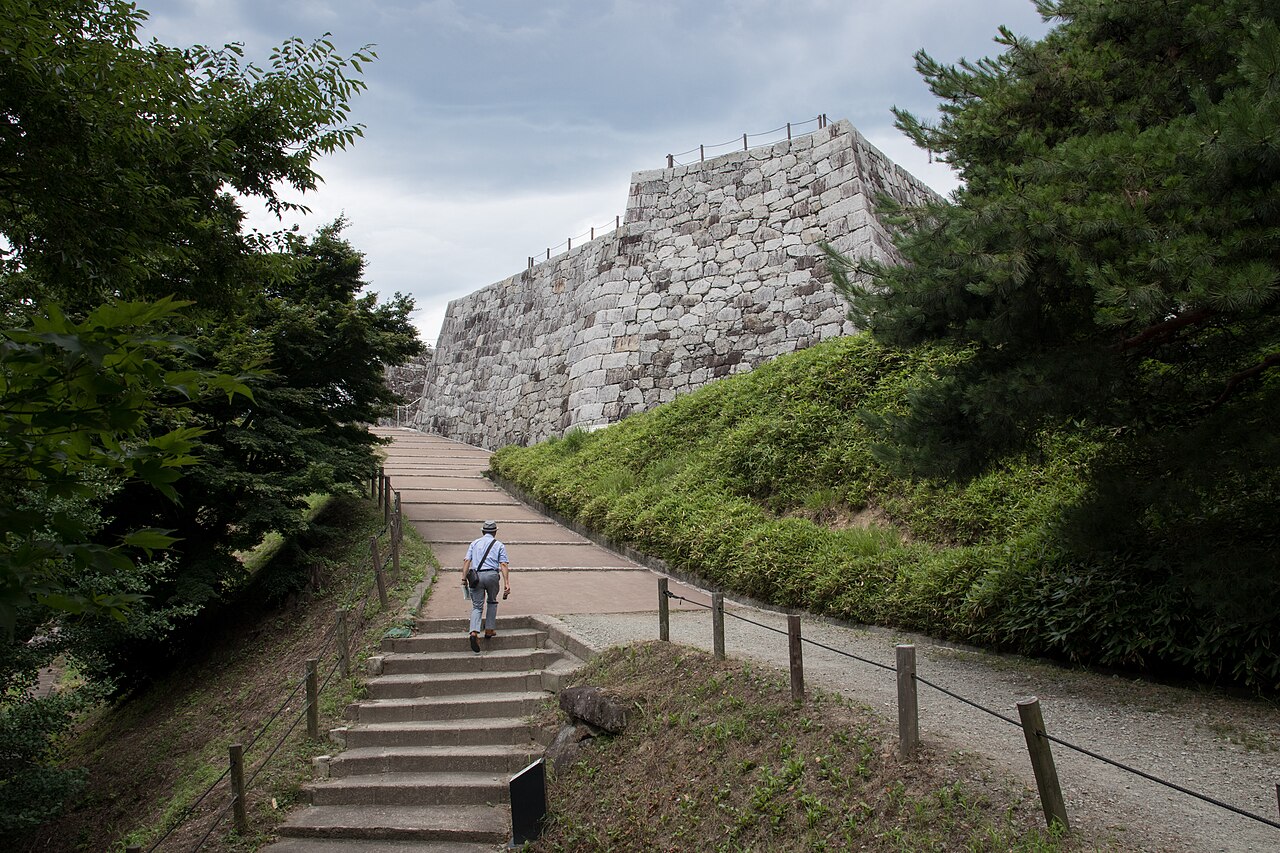 A long path with steps climbing beside a grassy bank to a massive stacked-stone castle wall under a cloudy sky, one visitor walking ahead