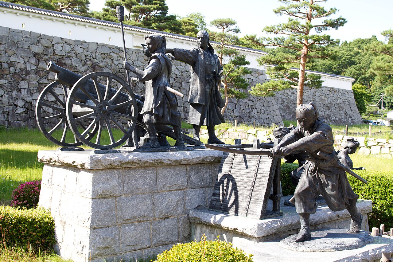 Bronze statues of young swordsmen and a boy loading a cannon on a stone pedestal, in front of a stacked-stone castle wall and white plastered wall
