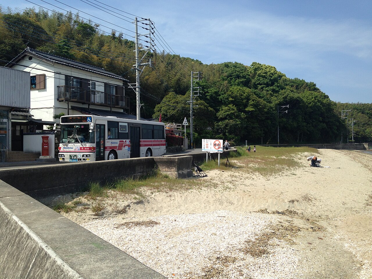 A red-and-white Nishitetsu bus on a coastal road beside a house, above a sandy shell beach with a few people and wooded hills behind