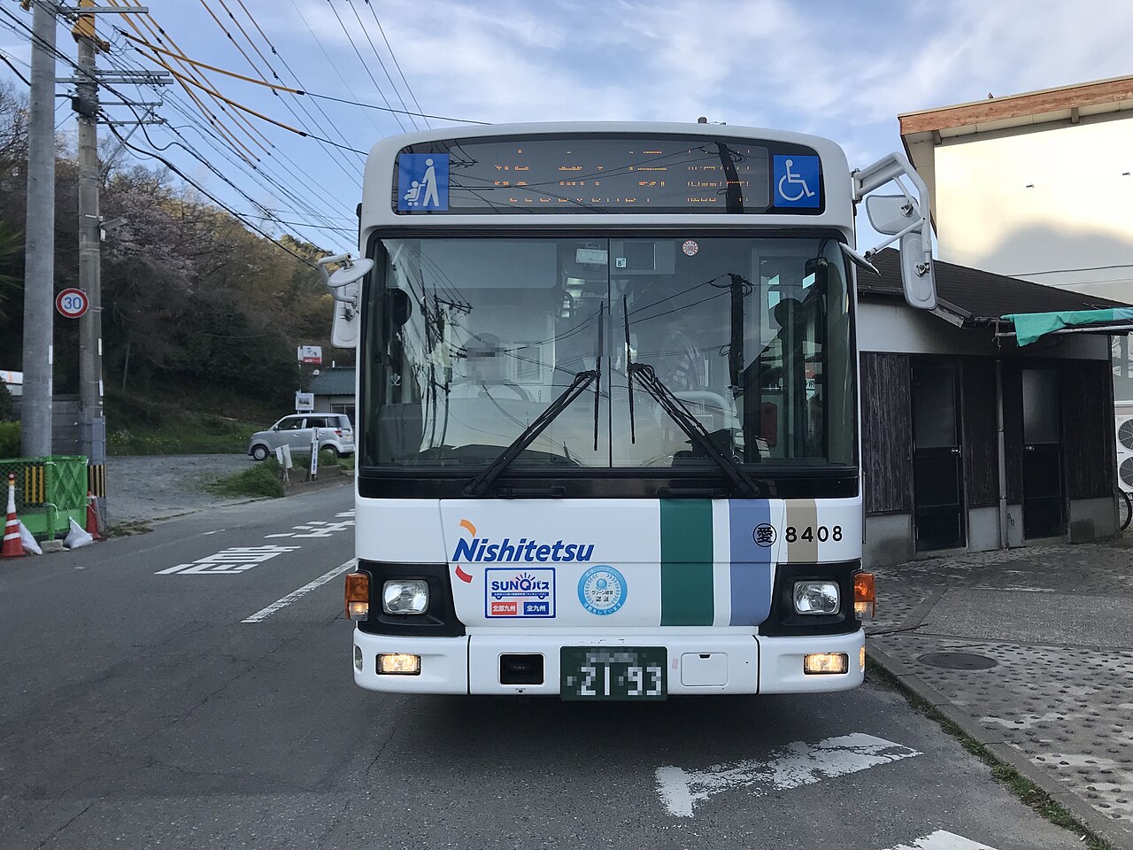 A white Nishitetsu bus with a green and blue stripe on its front, stopped on a narrow island road beside a small building