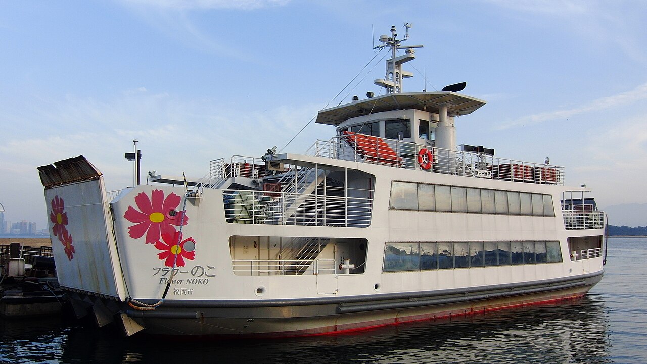A white two-deck ferry with large pink cosmos flowers painted on its side and bow ramp, moored at a pier under a pale blue sky