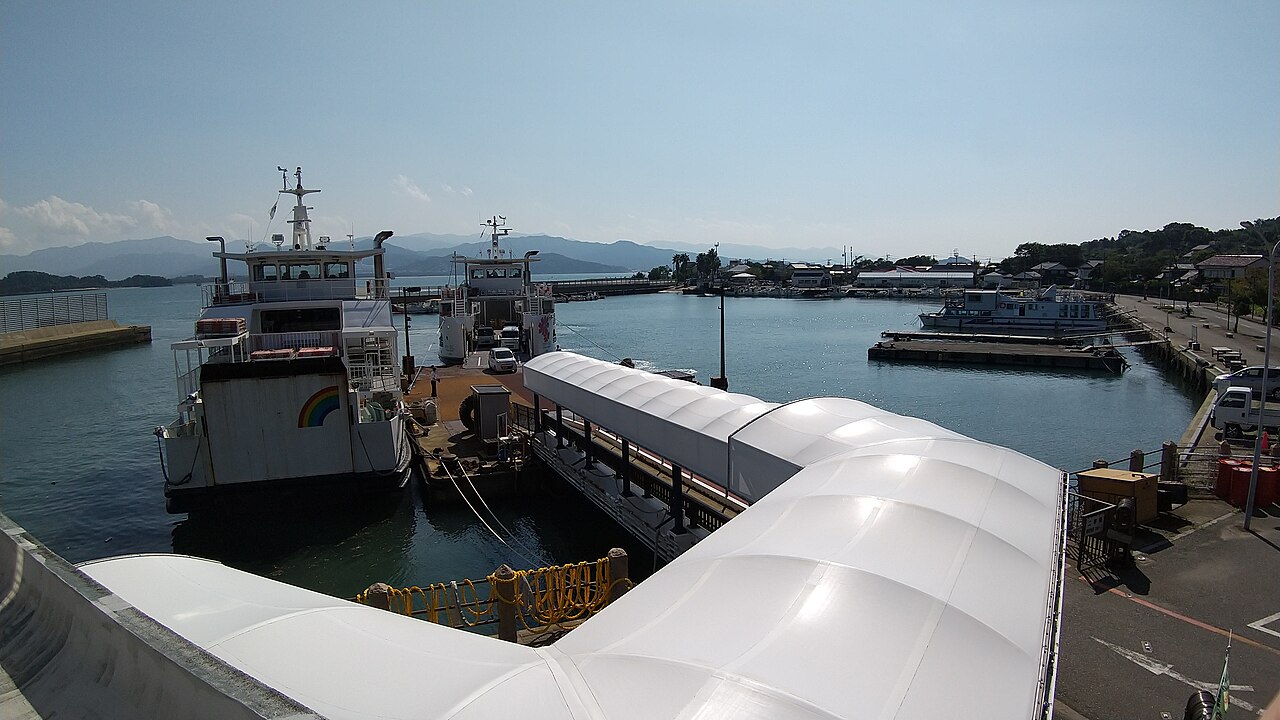 Two ferries docked at a pier with a white arched covered walkway in the foreground, with a small harbor and low hills behind