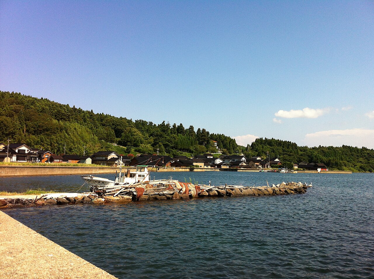 Small fishing boats beside a stone-block breakwater, with a row of black-tiled houses along the shore and a forested hill behind