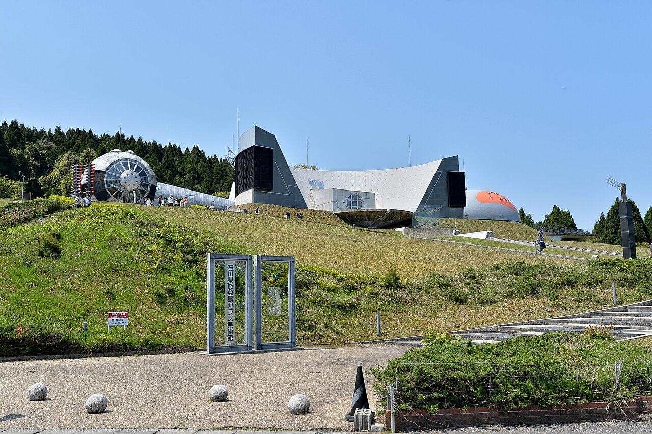 A silver angular building with a dome stands on a hilltop, with a glass gate in the foreground