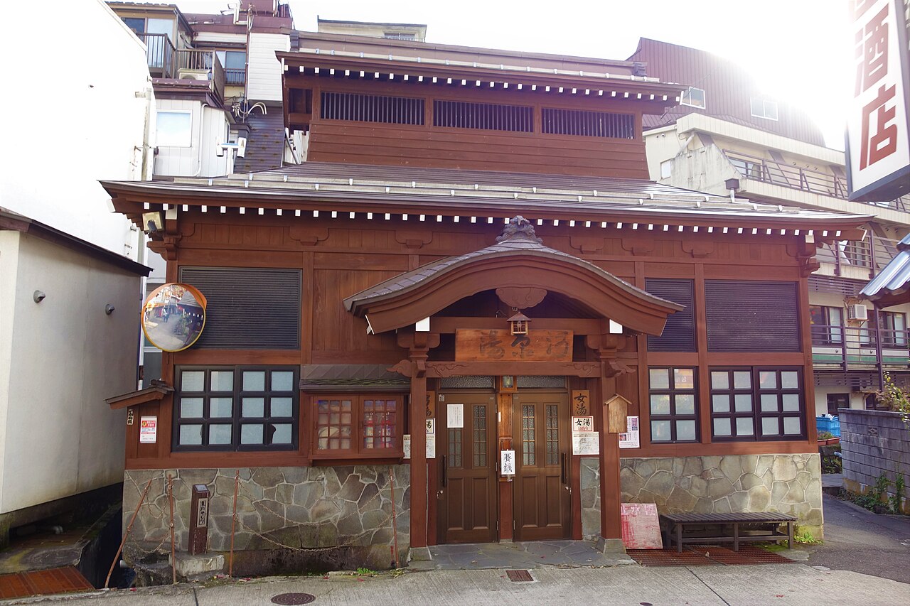 Exterior of a small wooden public bath house in Nozawa Onsen village, with a tiled entrance roof and stone base