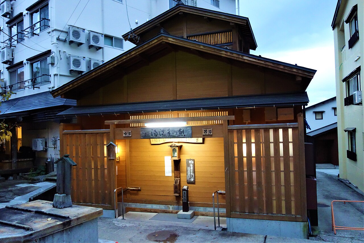 A small wooden public bath house in Nozawa Onsen lit warmly at dusk, beside a small stone basin