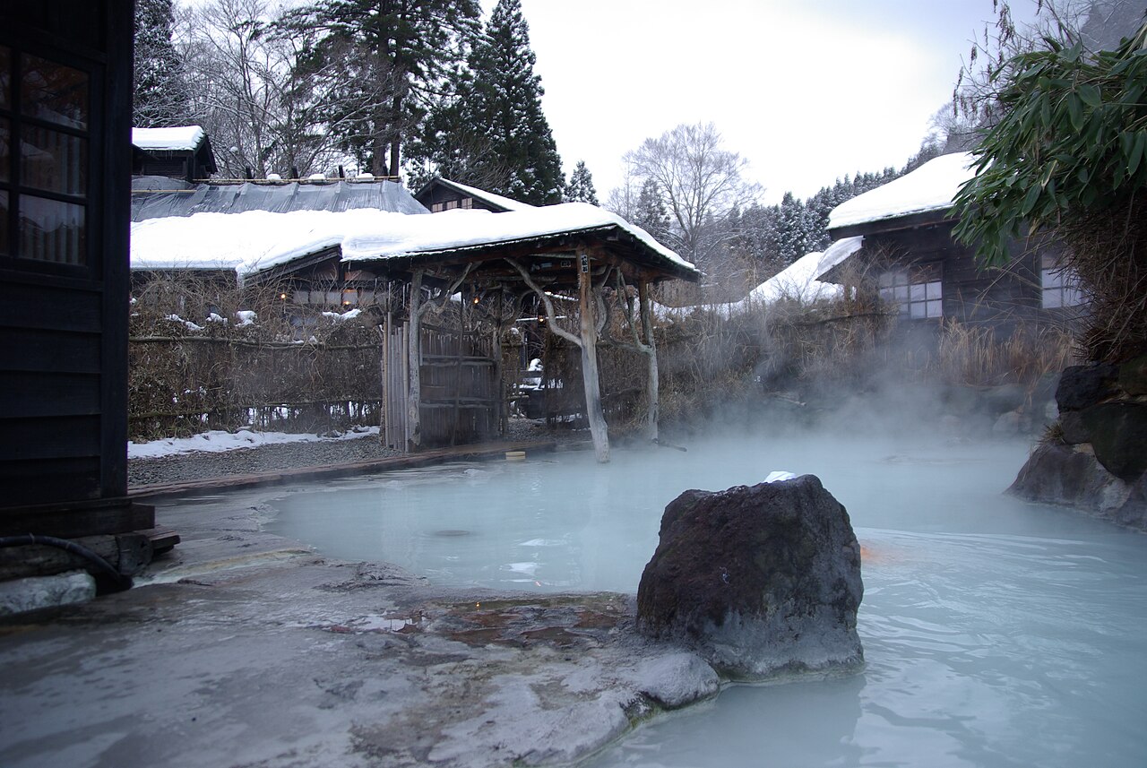 Steaming milky blue-white outdoor bath with a large rock at Tsurunoyu Onsen, snow on the roofs of the bathing huts behind