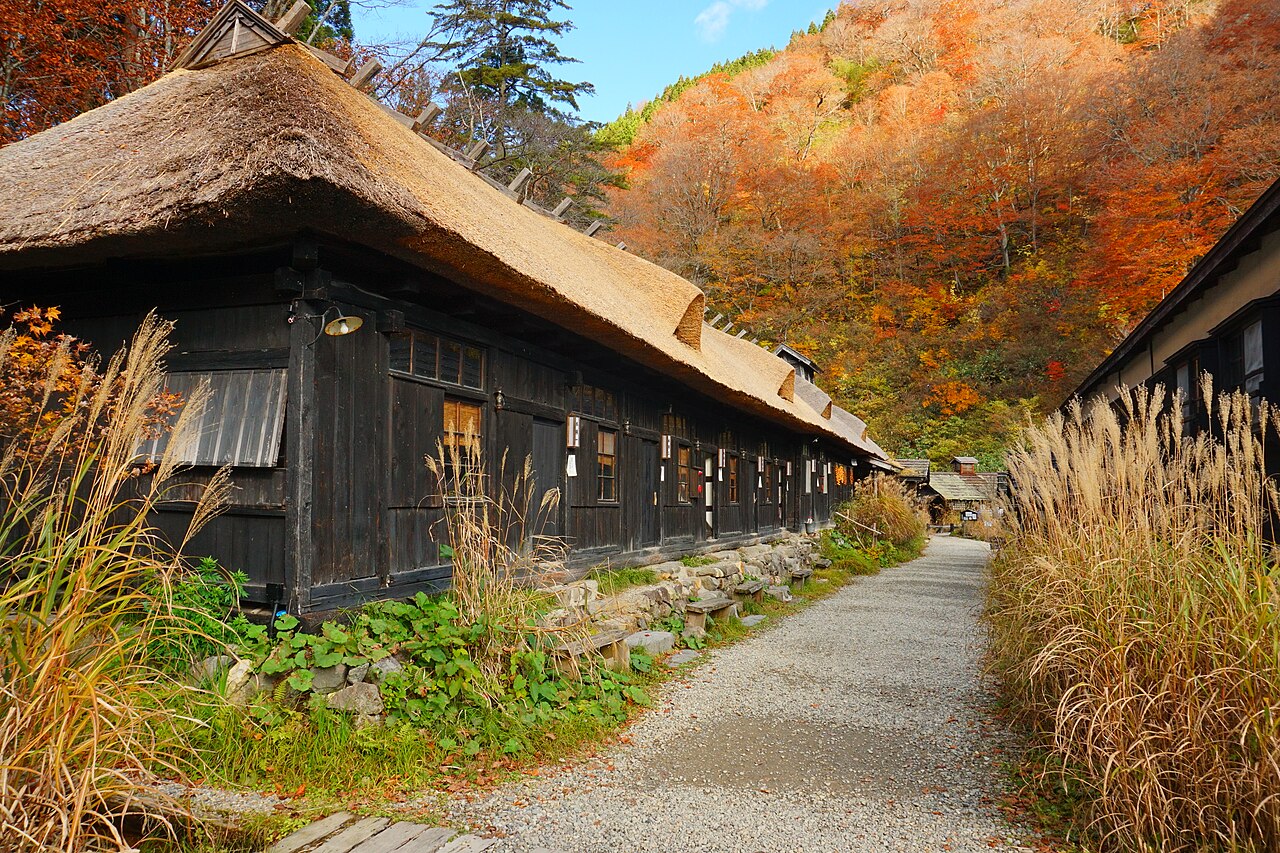 Long thatched-roof black timber bathhouse building at Tsurunoyu Onsen beside a gravel path, with pampas grass and autumn-coloured hillside behind