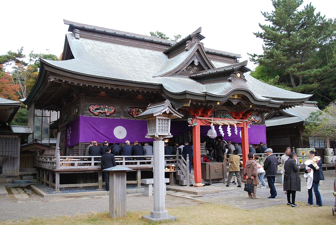 The Haiden worship hall of Oarai Isosaki Shrine with a tiled roof, red railings and a purple curtain, and a small group of worshippers in front