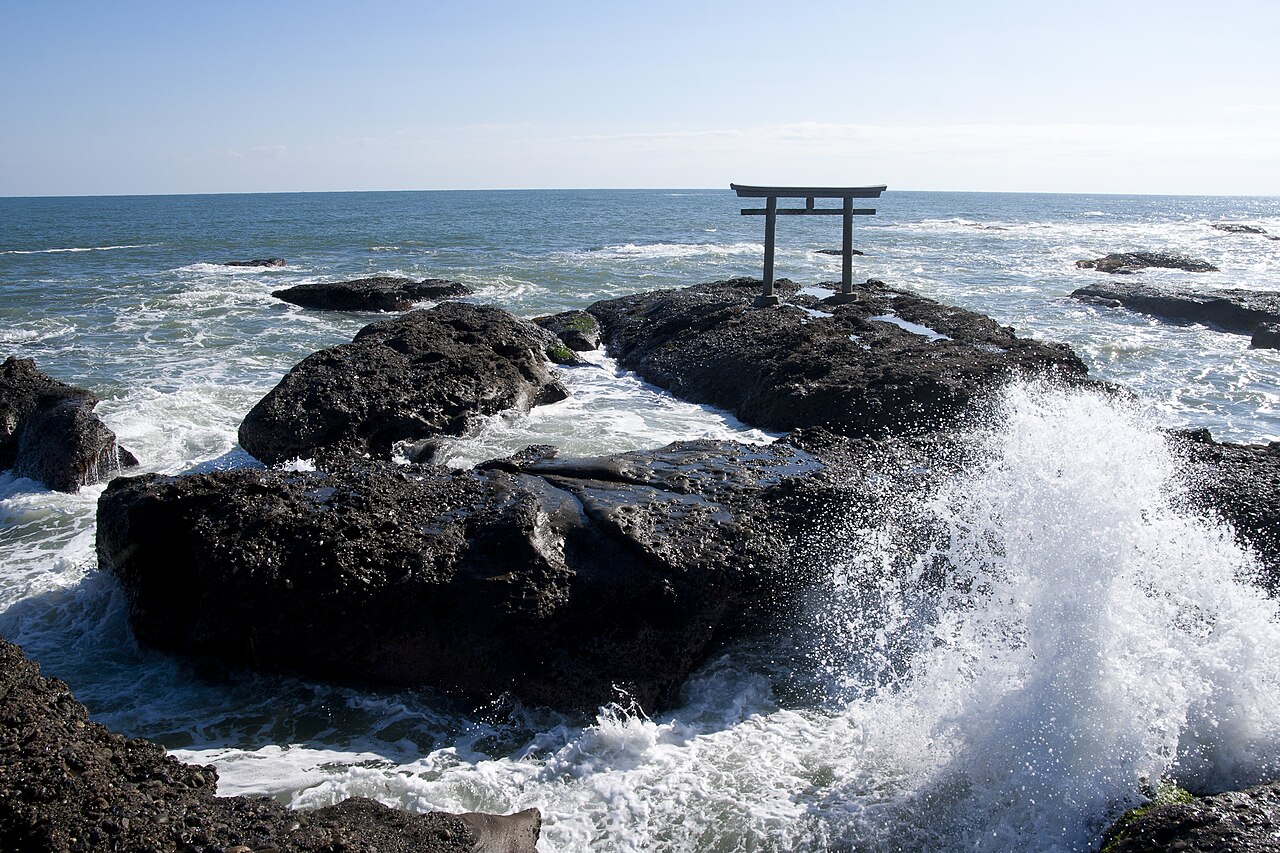 A small torii gate on a rocky outcrop while a wave breaks over dark rocks in the foreground on the Oarai coast
