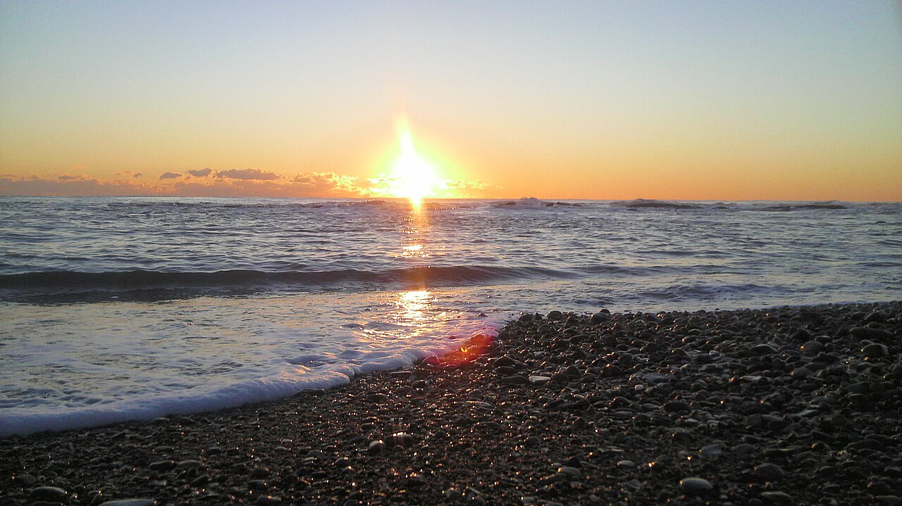 Sunrise on the horizon above a calm sea, reflecting on the water, with a pebbly beach in the foreground at Oarai Sun Beach