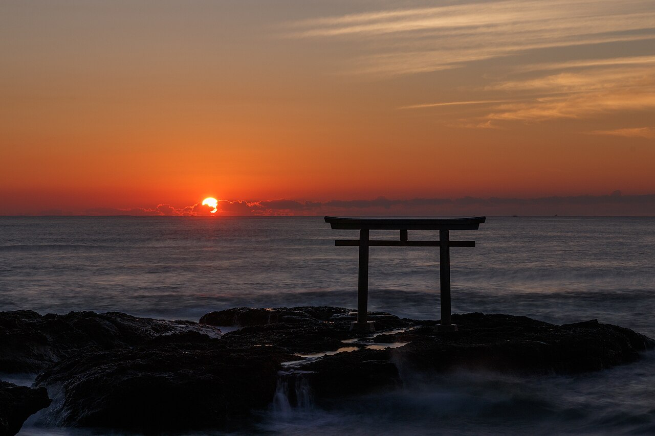 A red-orange sunrise over the Pacific with the Kamiiso torii gate standing on dark rocks at the water line on the Oarai coast