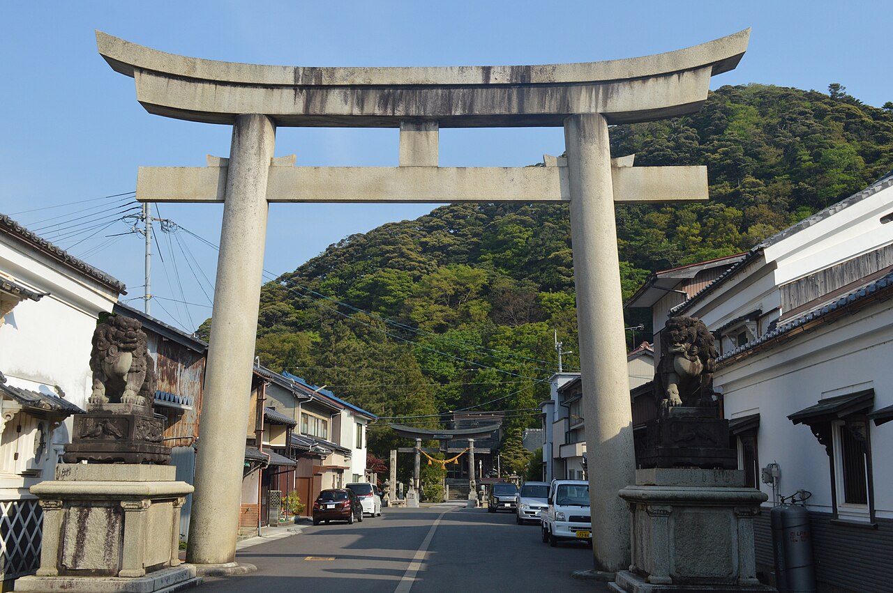 Large stone torii gate and a pair of stone guardian dogs on a street of townhouses leading to the Hachiman Shrine in Obama, with wooded hills behind