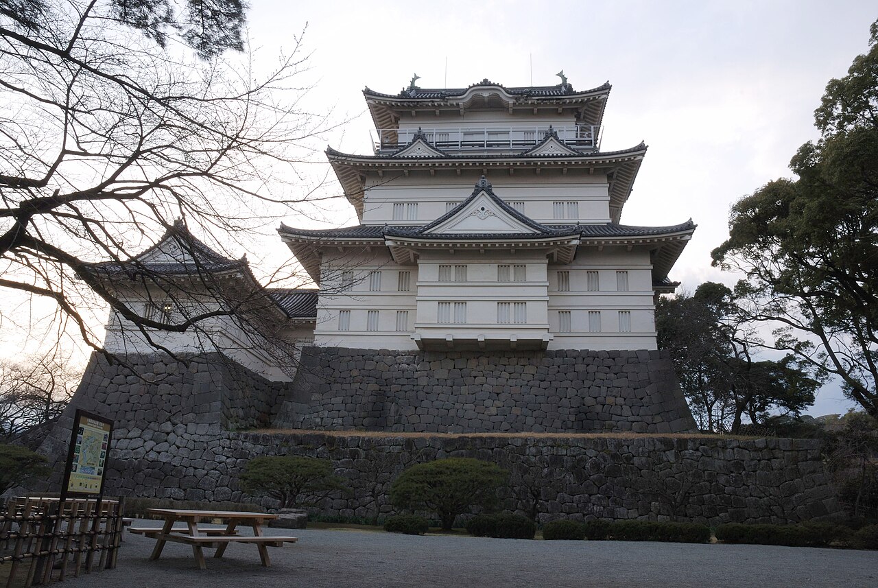 The white main keep of Odawara Castle on its stone base, with bare winter branches in front, a bench and an information board.
