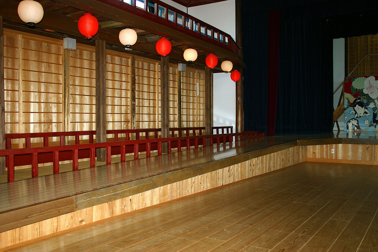 The wooden stage and box seating inside the Odeon-za theater in Wakimachi, hung with red and white paper lanterns beside a painted kabuki backdrop.