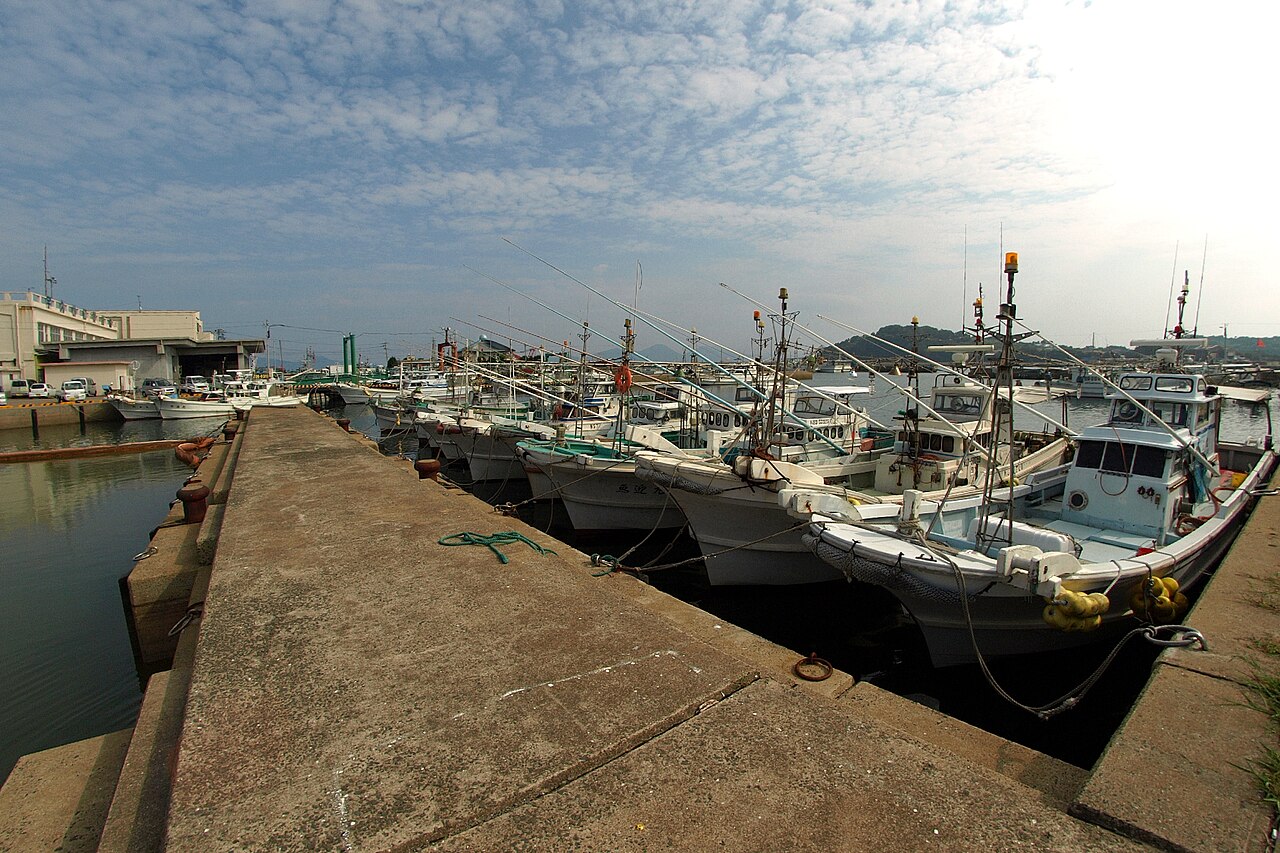 Rows of small fishing boats with fishing rods and masts are moored along a concrete quay under a wide sky of scattered clouds.