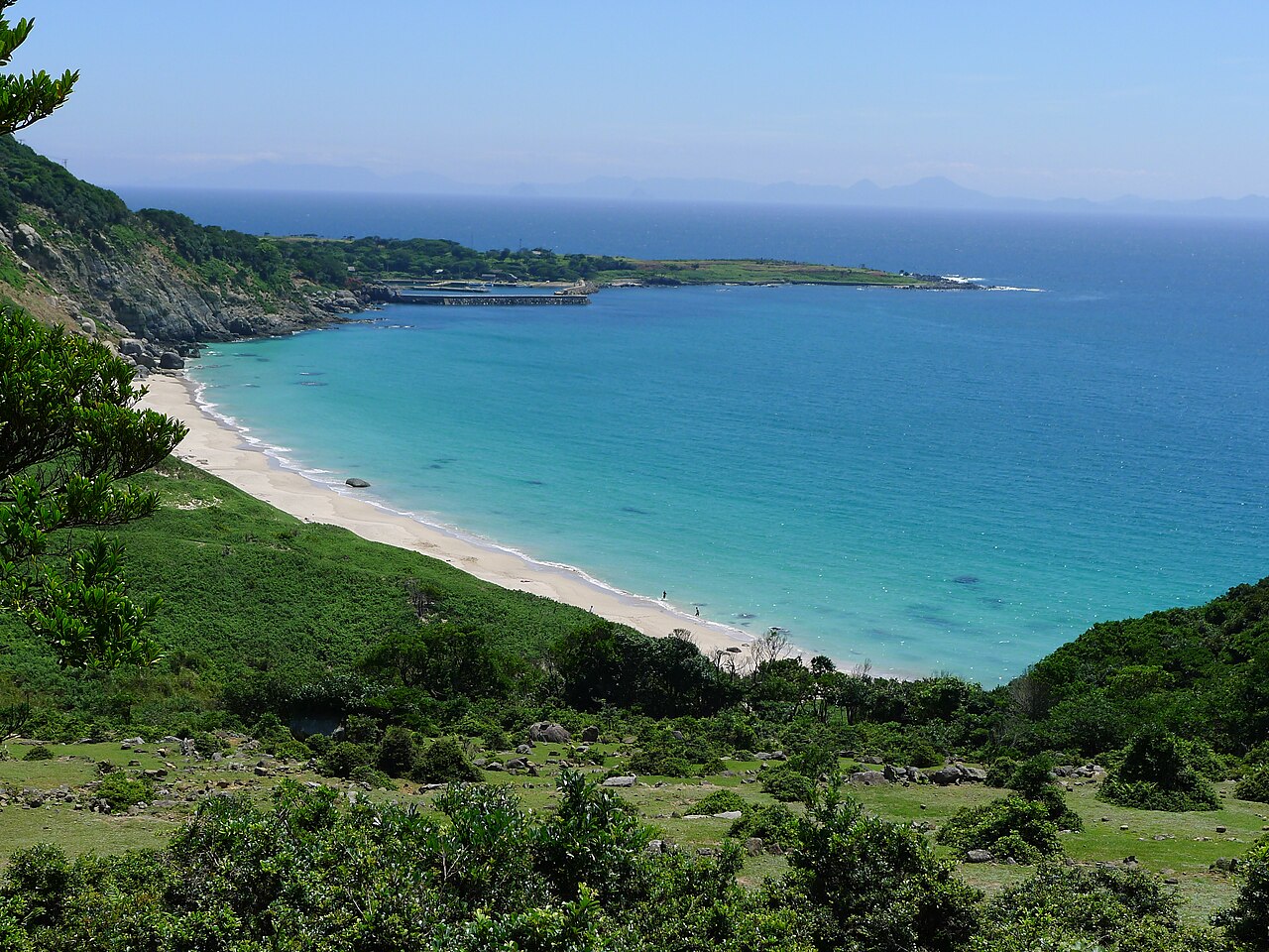 A long white-sand beach curves along turquoise water below green hillsides, with a small harbour breakwater on the far headland and hazy islands on the horizon.