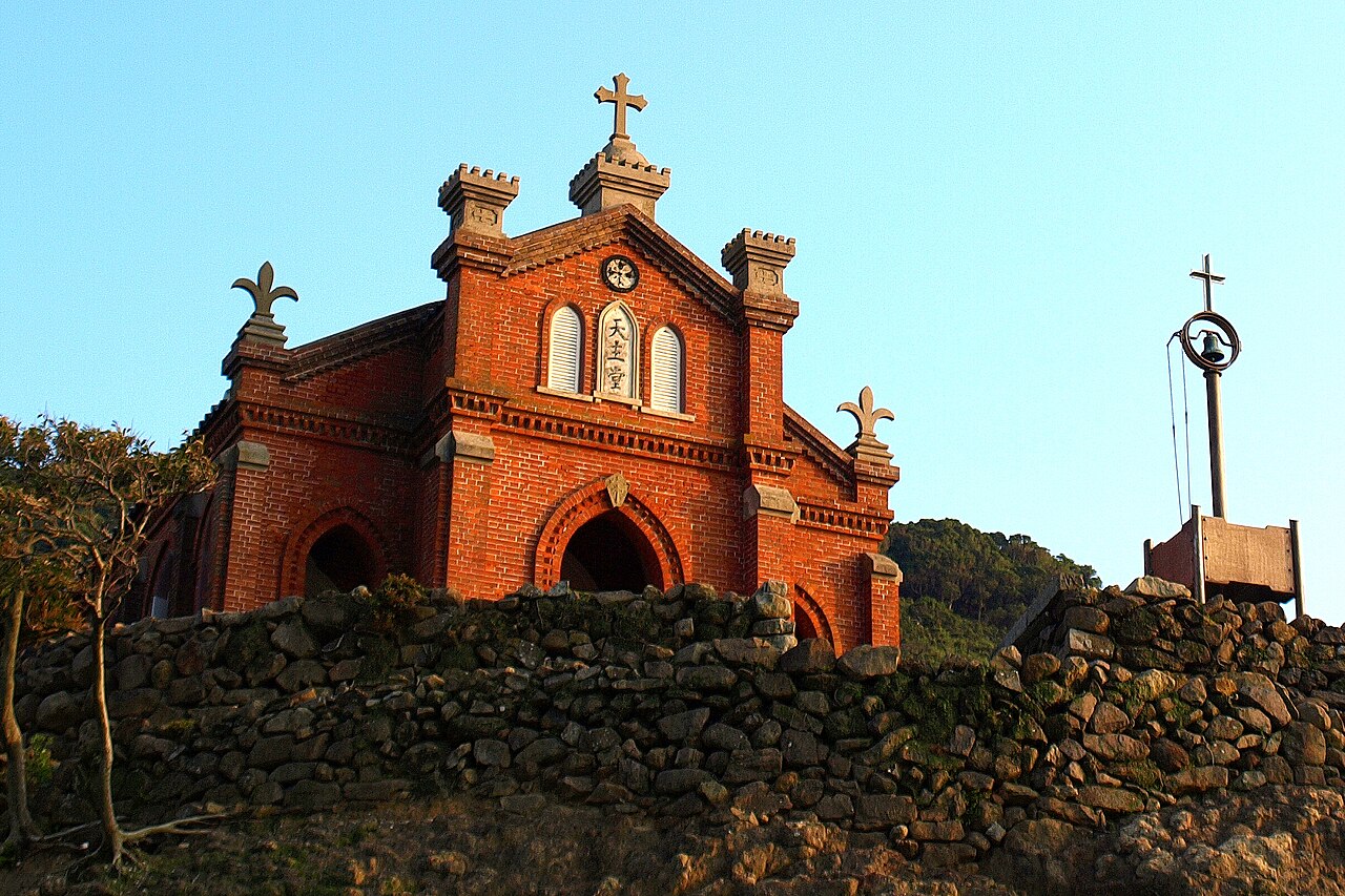 A red-brick church with a stepped gable, a cross on top and arched windows stands behind a dry-stone wall under a clear blue sky, with a small bell frame on a post at the right.