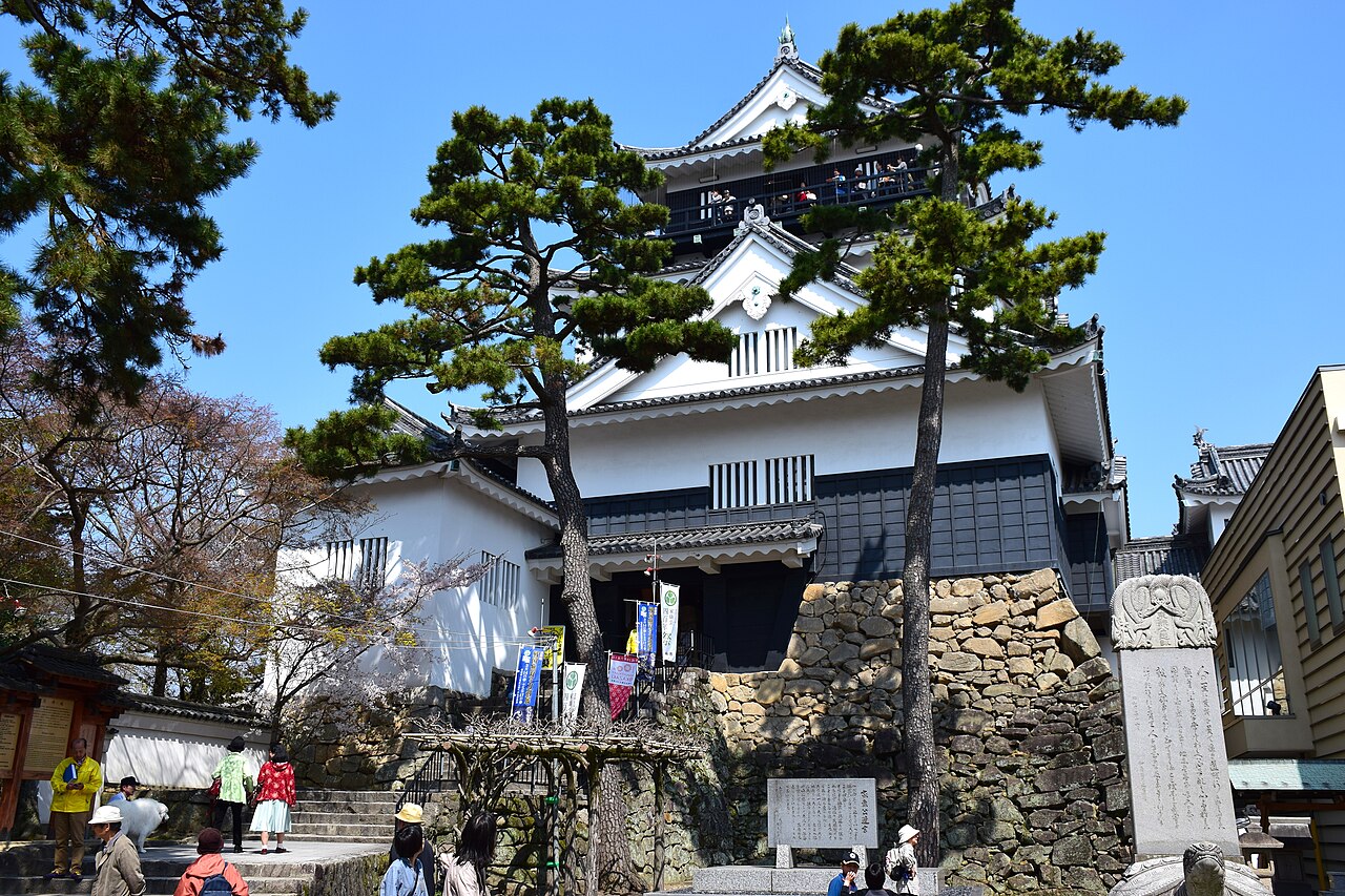 Okazaki Castle keep with white walls and dark roofs rising behind pine trees above a stone-walled base, with visitors on the steps below
