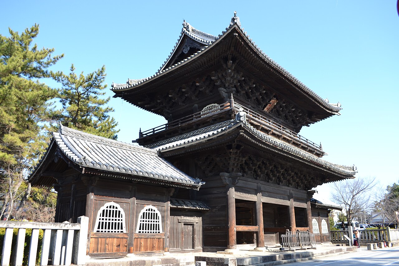 A two-storey wooden temple gate with layered tiled roofs and carved brackets under a clear blue sky at Daijuji Temple