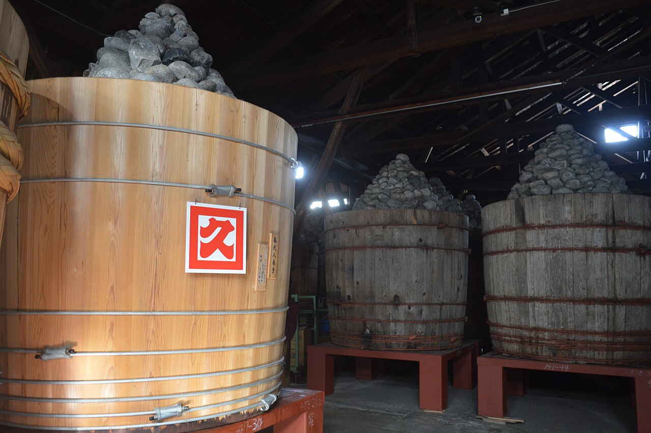 Large wooden barrels stand inside a miso storehouse, each topped with a heaped pile of weighting stones