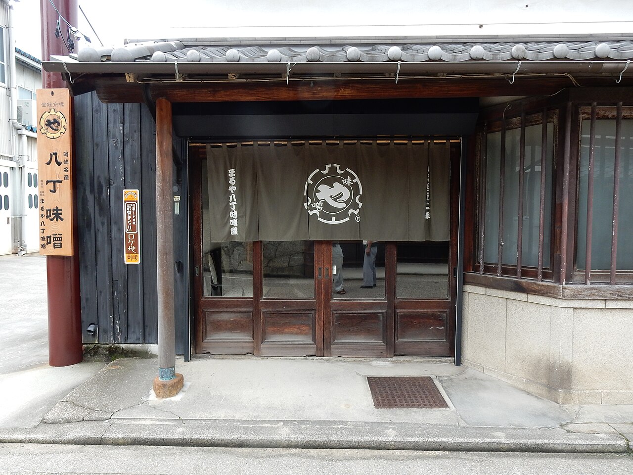 The front of a miso shop with a shop-name curtain at the door, a wooden lattice, black board walls and a tiled eave