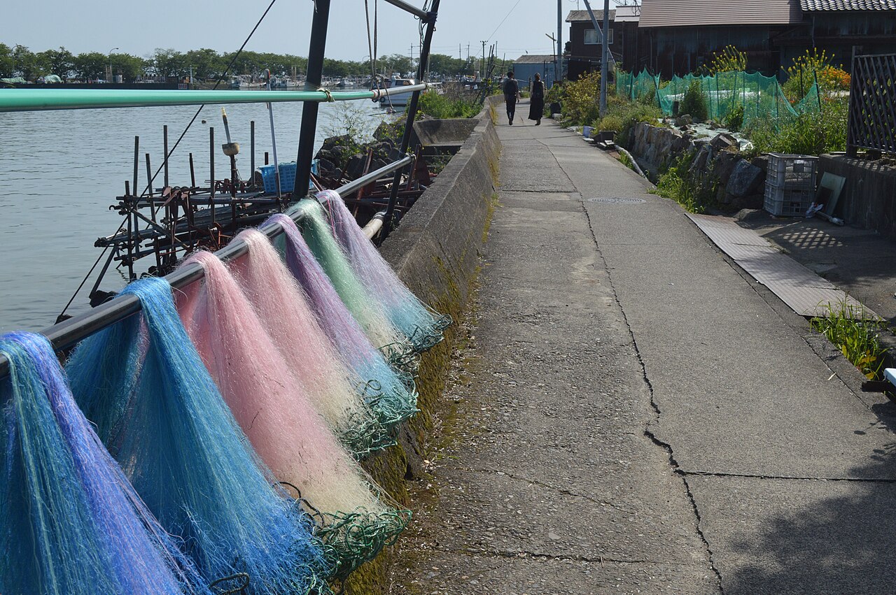 Pink and blue fishing nets hung along a cracked concrete quay path beside moored boats on Okishima.