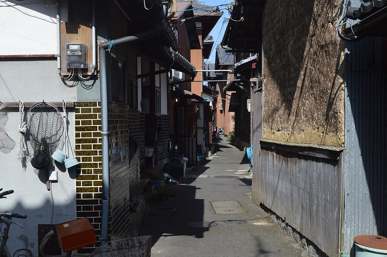 A narrow alley between weathered houses on Okishima with fishing gear hanging by the doors.