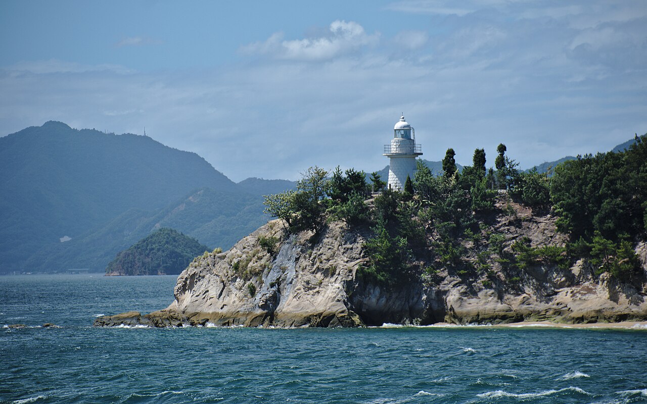 A small white lighthouse stands on a wooded rocky headland above choppy blue water, with hills behind.