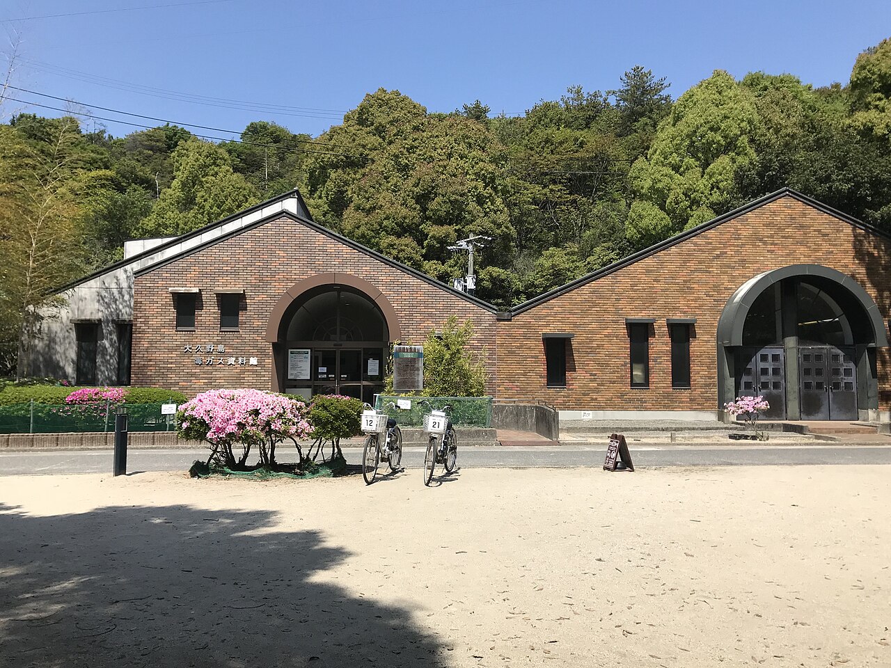 The low brick building of the Okunoshima Poison Gas Museum under a clear sky, with bicycles parked in the sandy square in front.