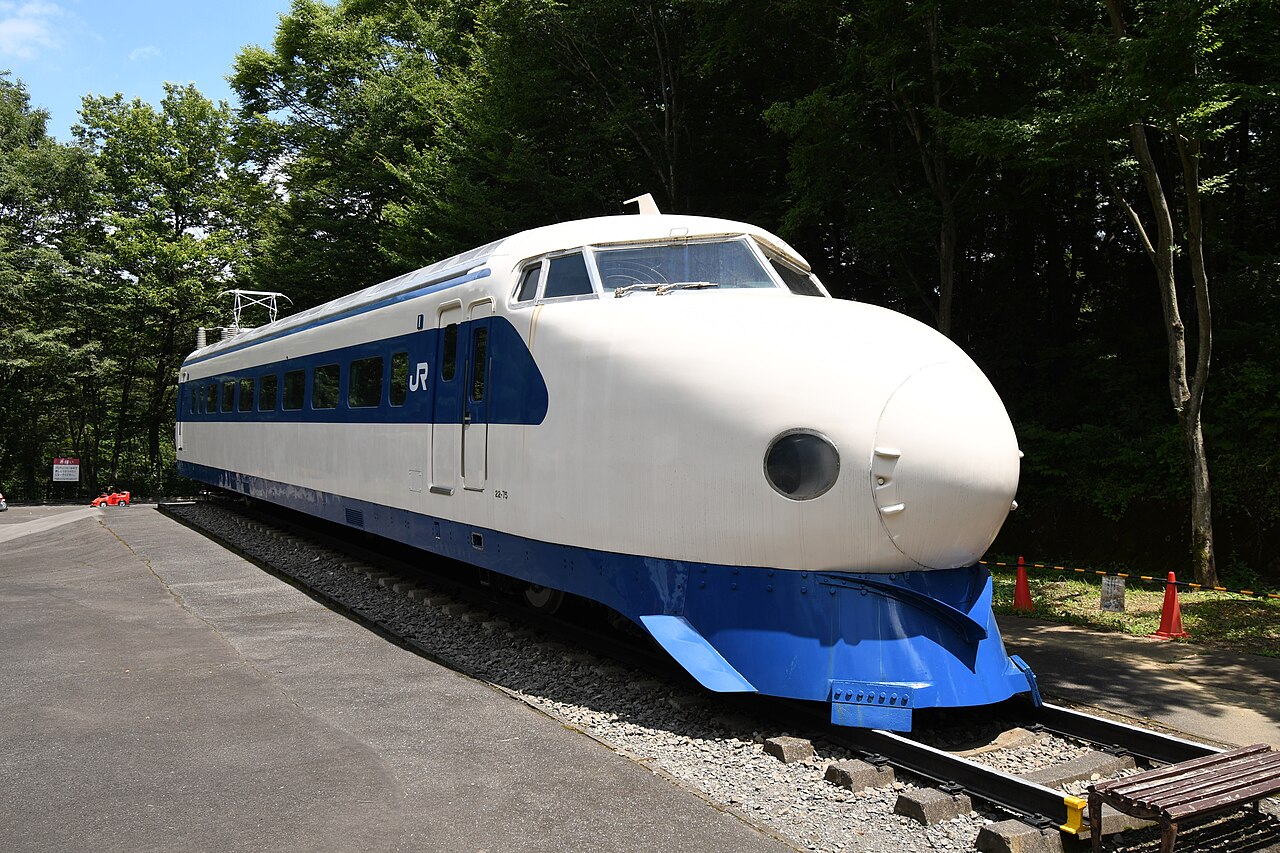 A preserved white and blue Series Zero Shinkansen car stands on a short stretch of track among trees, seen from a front corner in bright daylight, with a bench at the right edge.