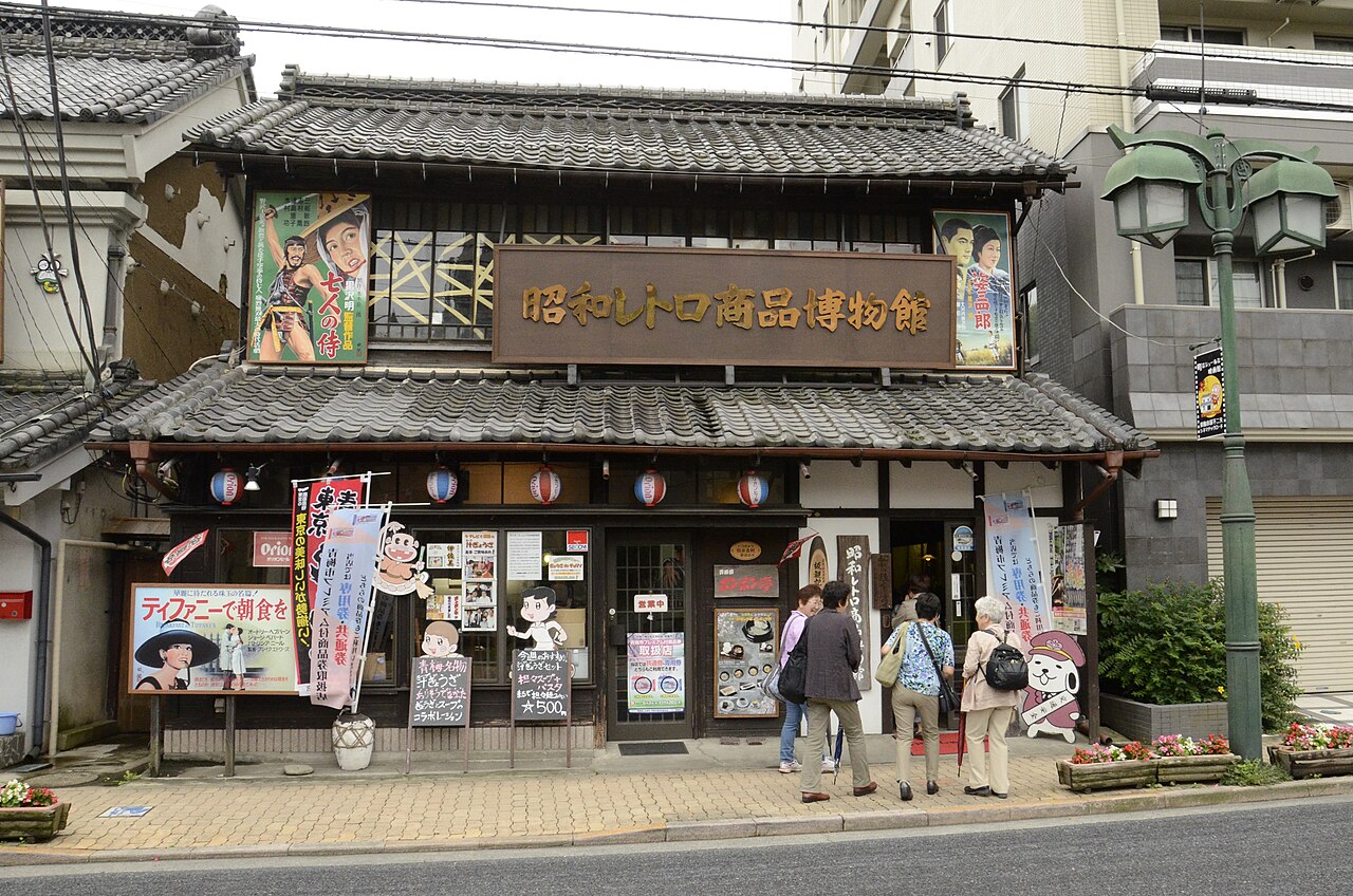 A two-storey old shop with a tiled roof and a large wooden signboard for the Showa Retro packaging museum, hung with vintage film posters and paper lanterns, with visitors at the entrance and a green lamppost at the right.