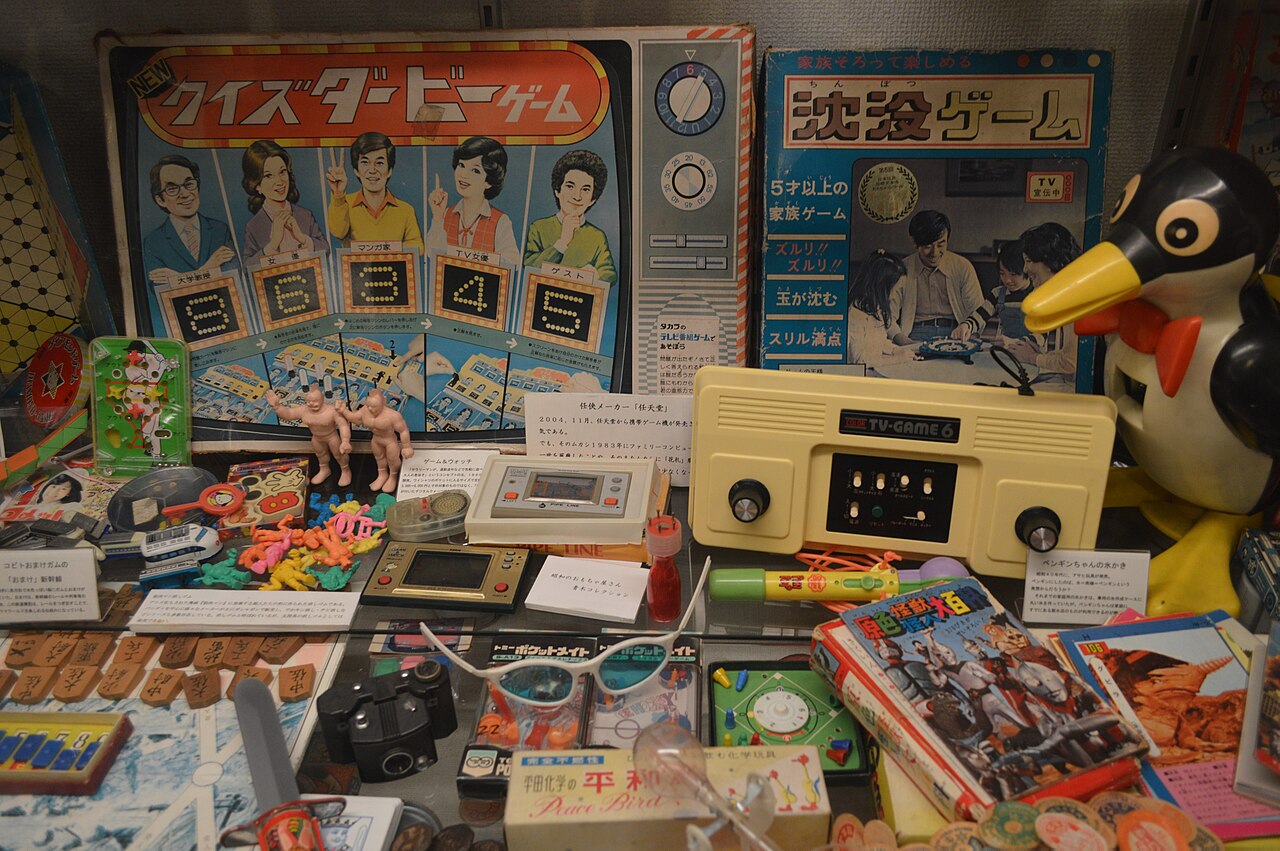 A glass display case crowded with vintage toys and games, including a TV quiz board game box, a cream-coloured TV Game Six console, handheld LCD games, small plastic figures and a black-and-yellow penguin toy.