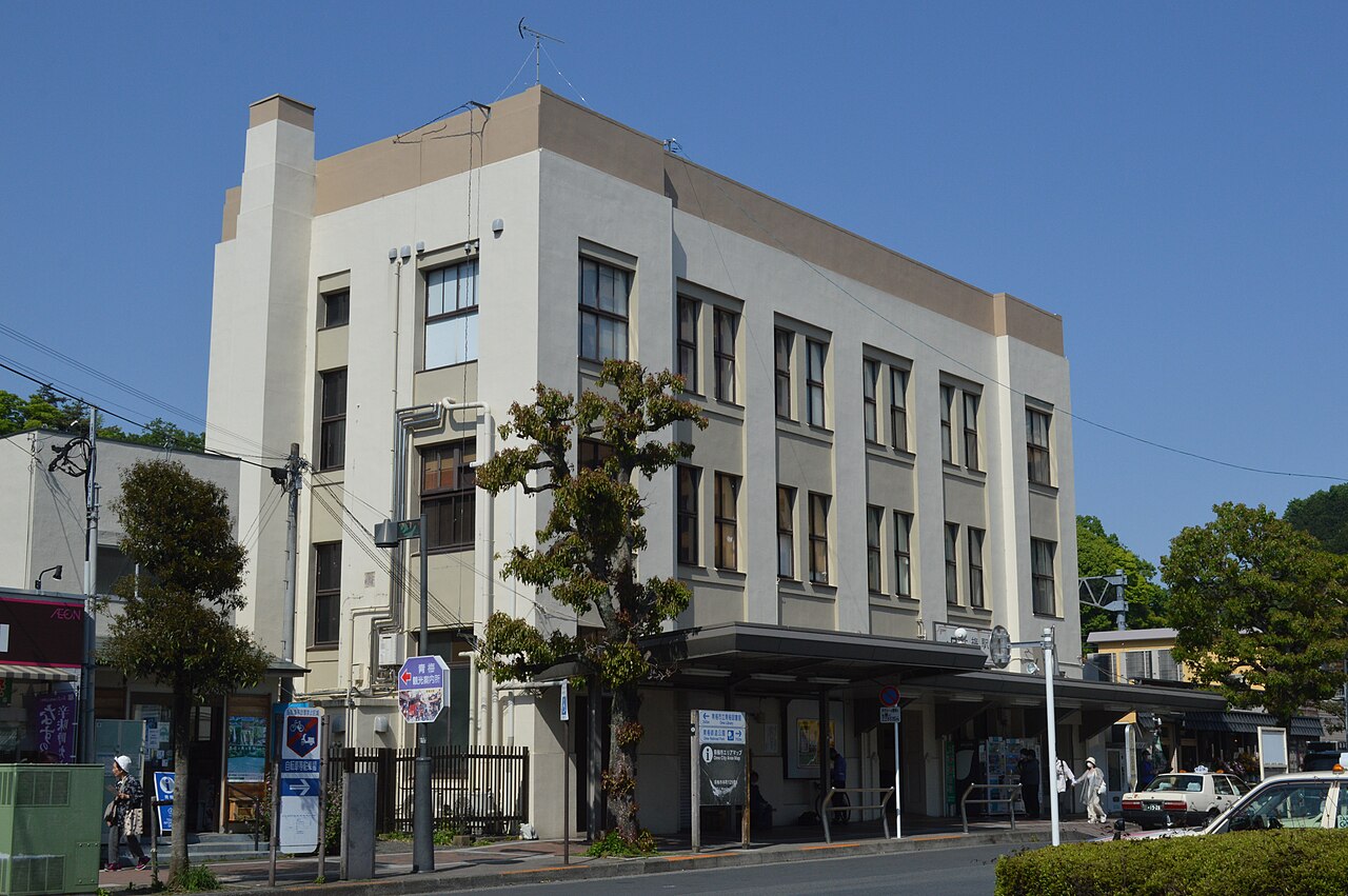 A cream-coloured three-storey station building with a flat roof and tall windows stands beside a bus stop and street signs under a clear blue sky, with a few people and a taxi at the right.