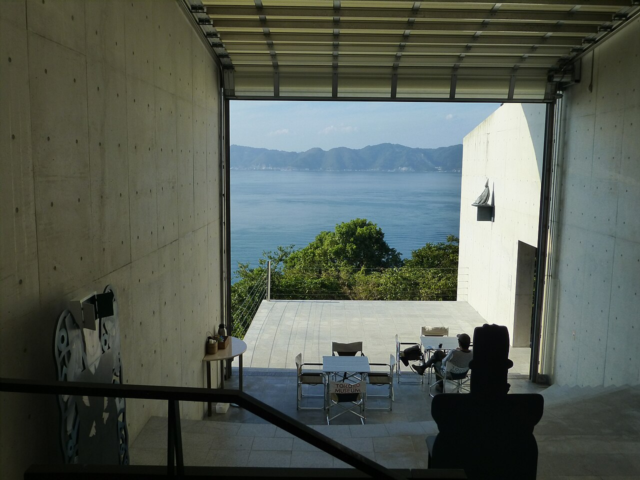 A concrete exhibition room with an opening that frames a blue bay, with chairs on a terrace