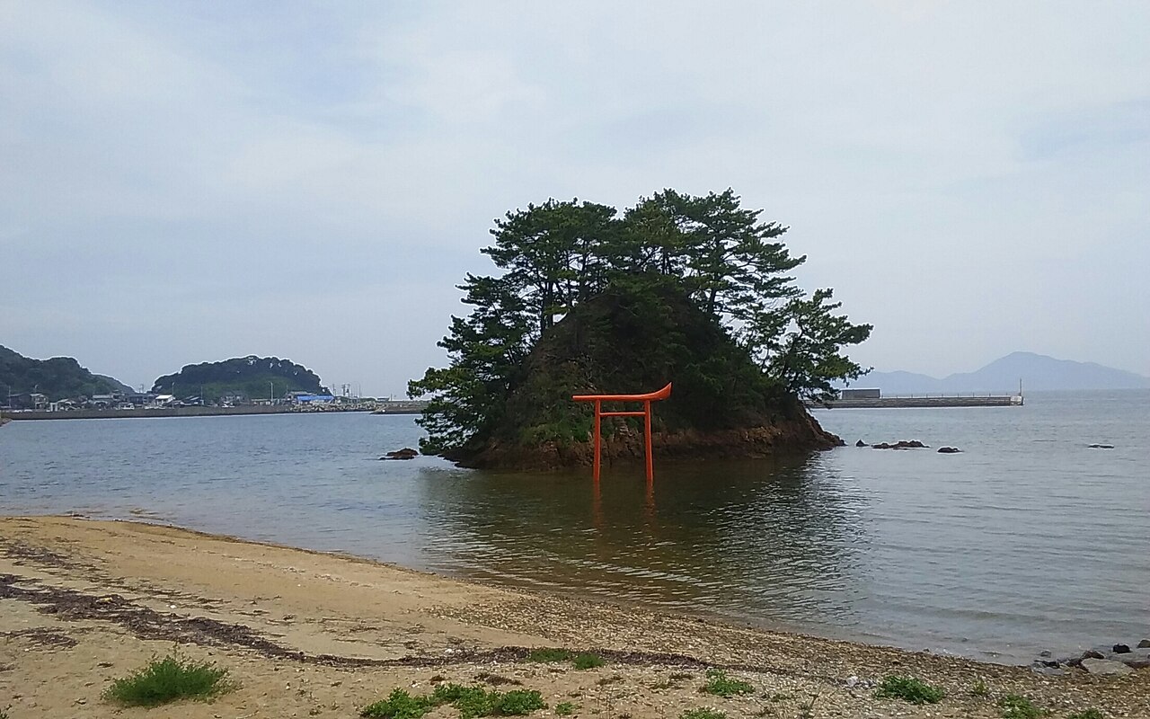 A small rocky islet crowned with pine trees off a beach, with a vermilion torii gate standing in the shallows in front of it, under a cloudy sky with a harbor and hills in the distance