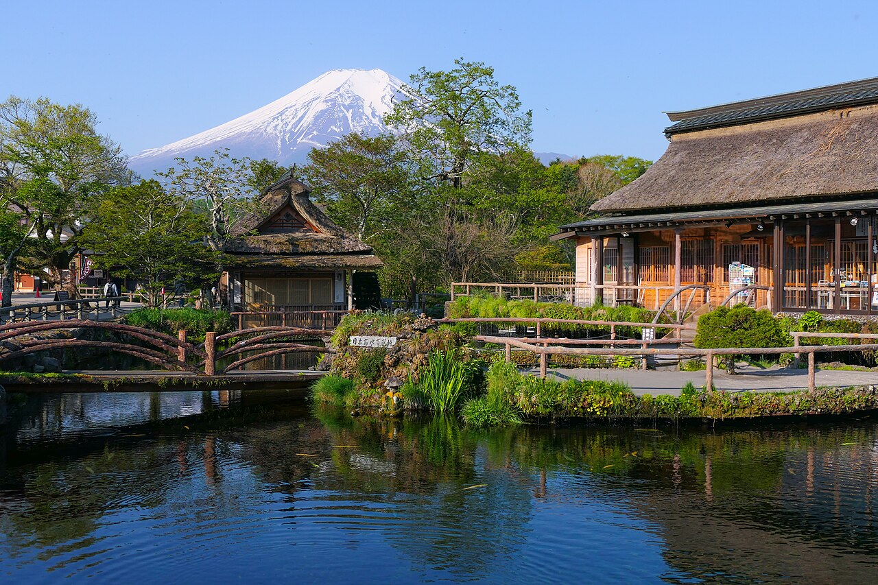 Mount Fuji rising over the wooden footbridge and thatched teahouse beside Naka Pond at Oshino Hakkai, with trees and still water in the foreground