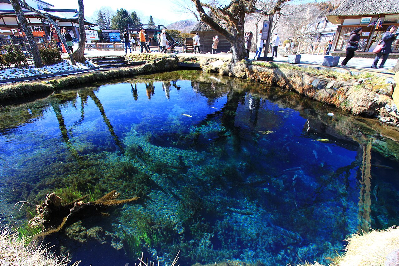 The deep blue, crystal-clear water of Waku Pond at Oshino Hakkai, with water plants on the bed and a few visitors and a thatched building on the far bank