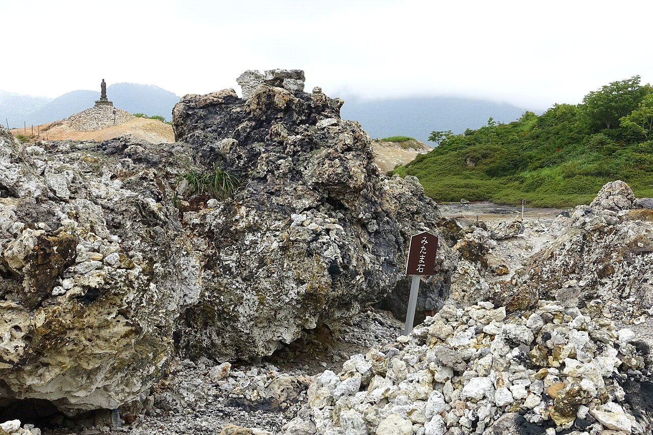 Large weathered volcanic rocks with a small marker sign and piles of white stones, with a stone figure behind